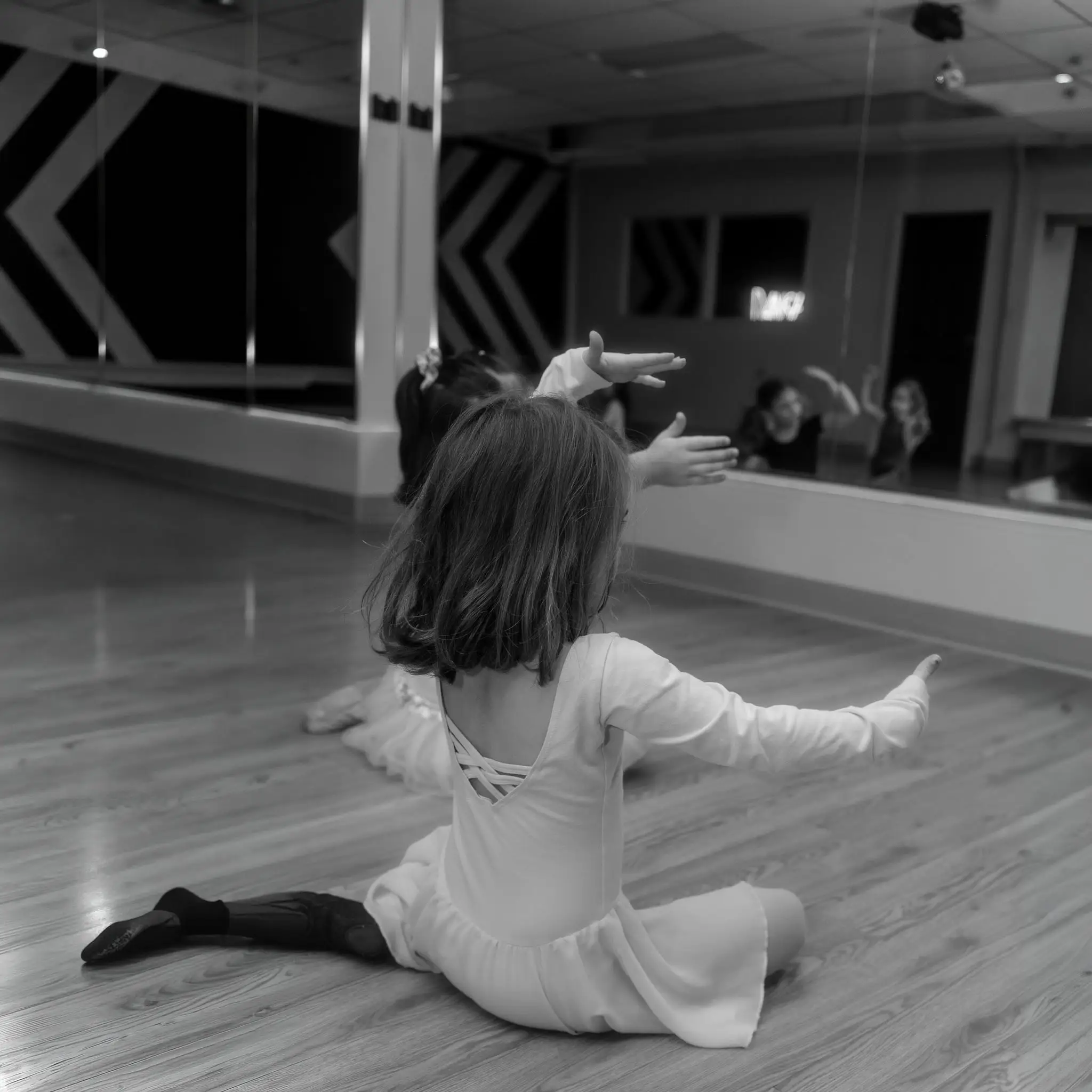Petite ballerine assise au sol, bras déployés face au grand miroir du studio