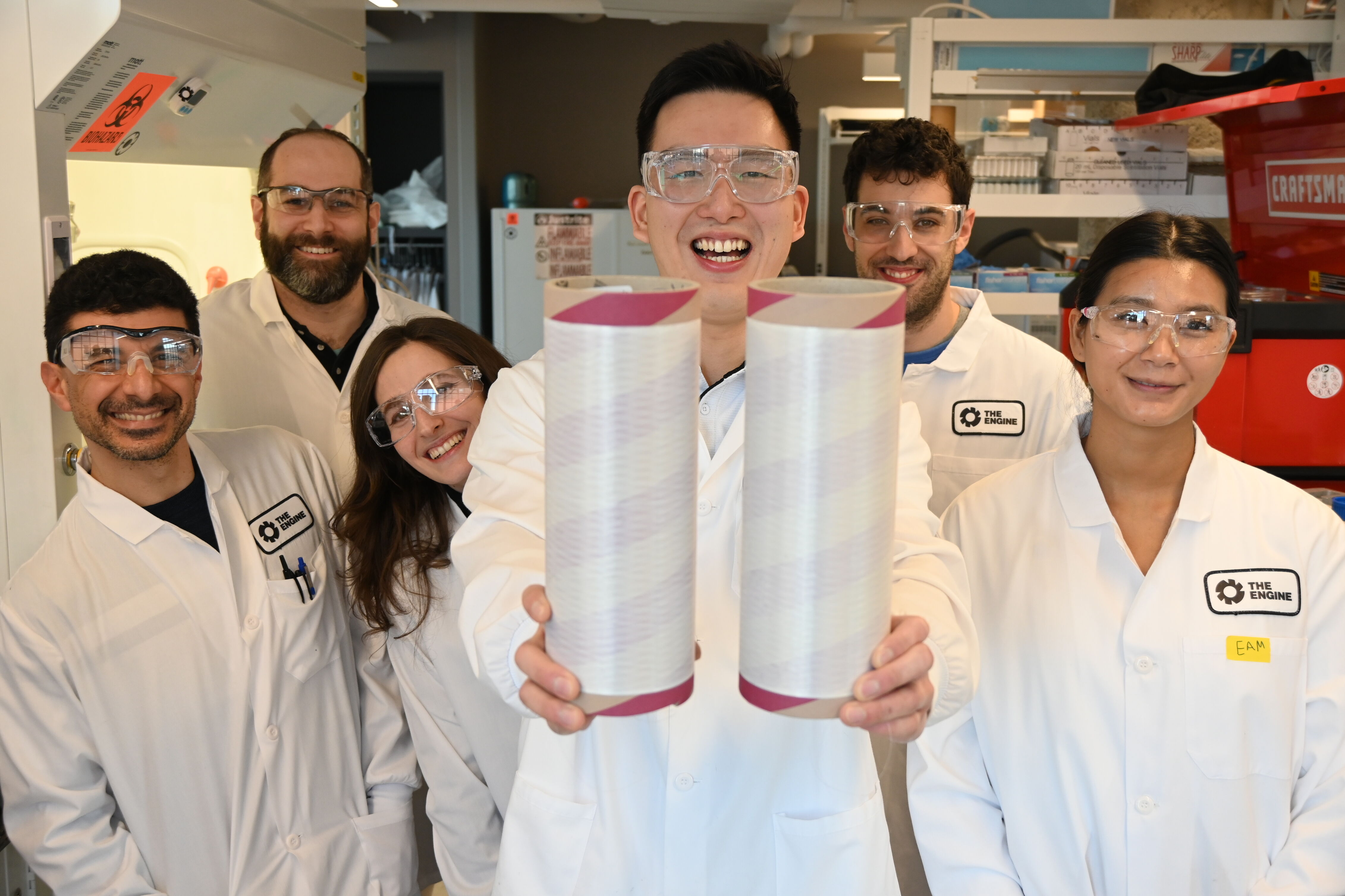 Six diverse scientists in lab coats and safety glasses smiling, one holding two cylindrical objects.
