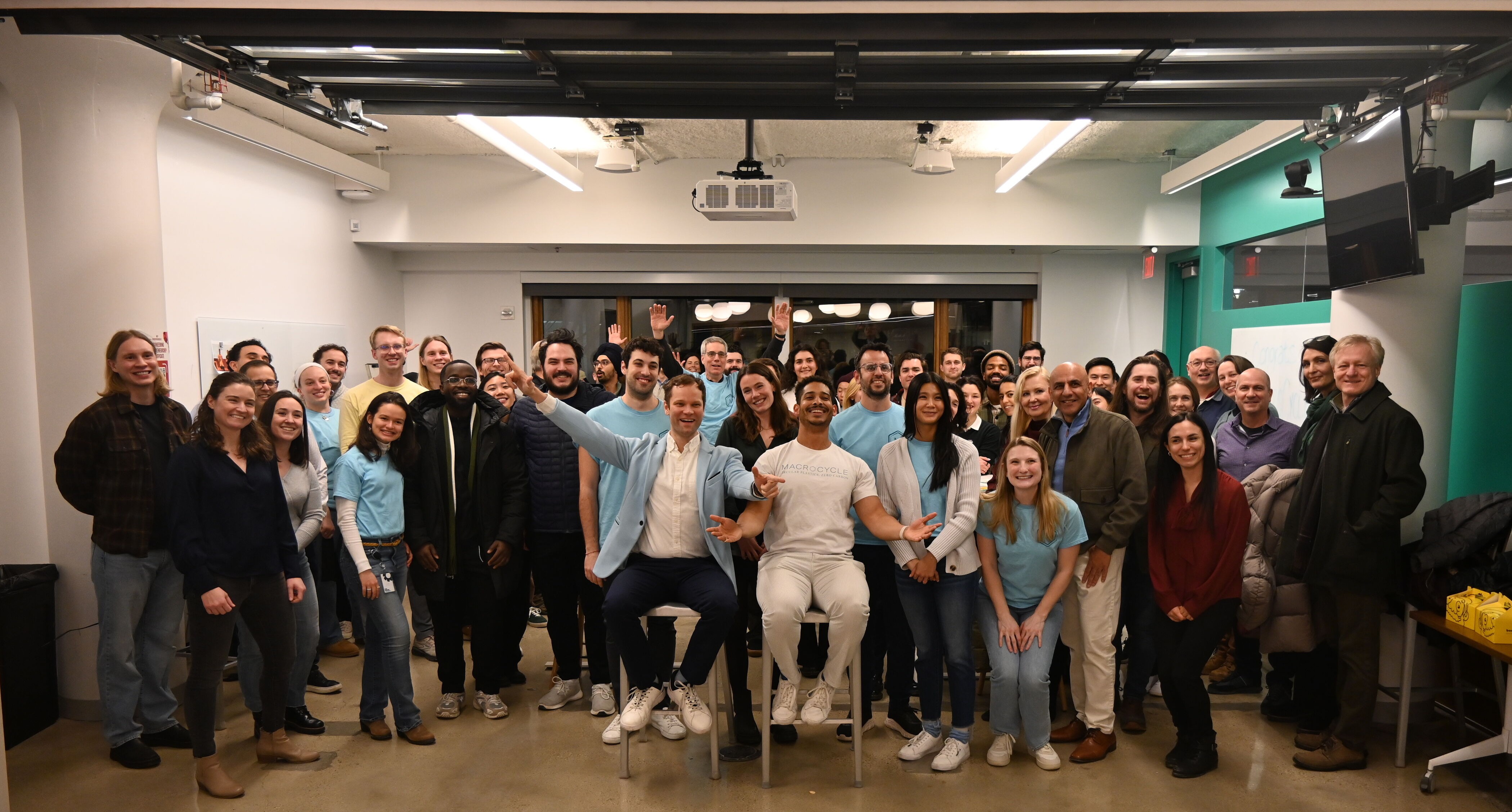 Large group of diverse people smiling and posing indoors, with two men seated in front raising arms.