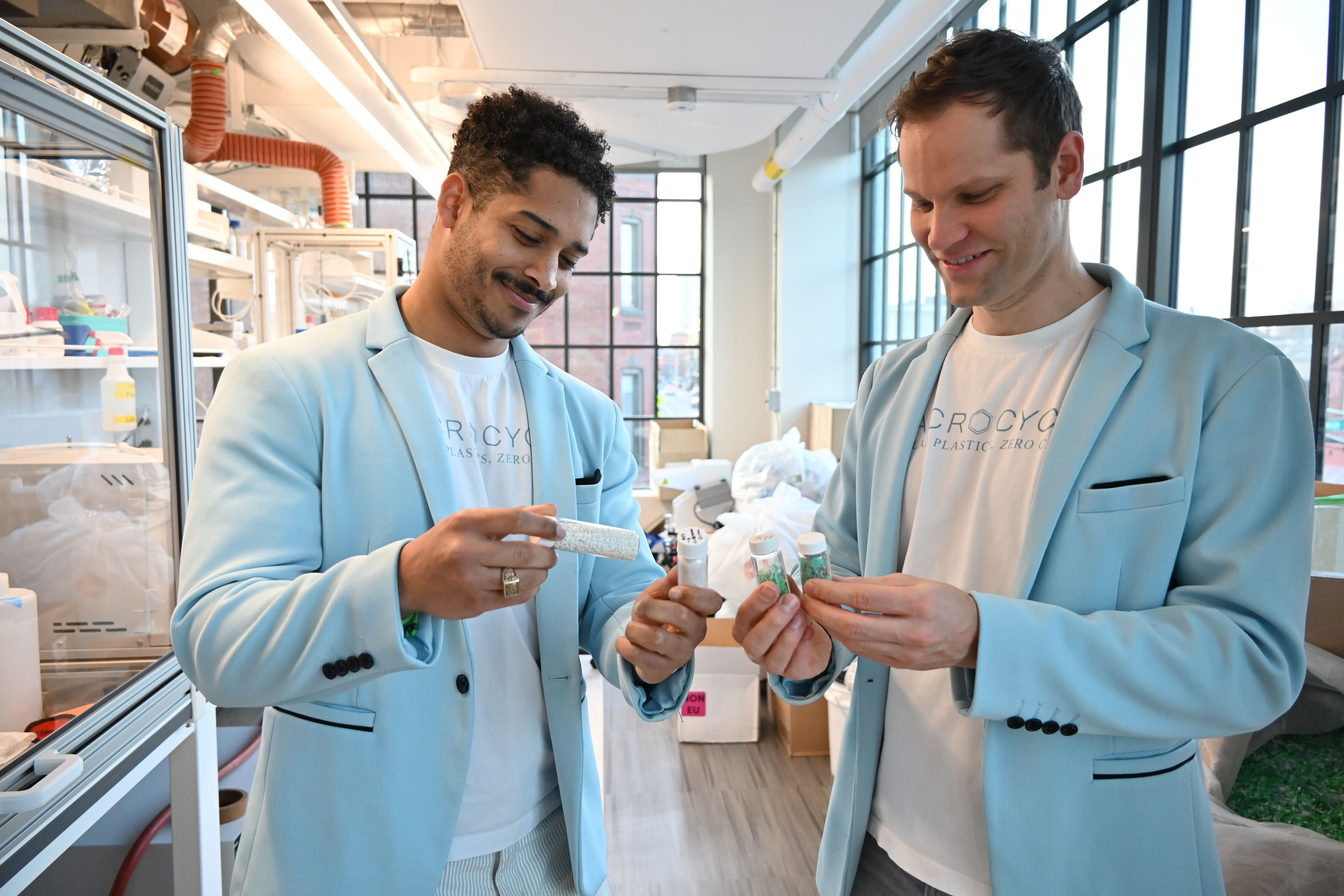 Two men in light blue jackets examining small containers in a bright, modern lab workspace.