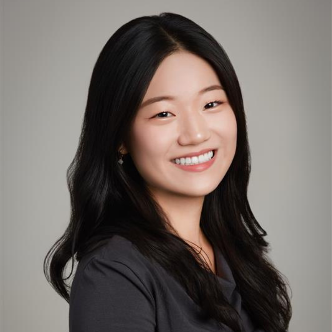 Smiling woman with long black hair wearing a dark top against a neutral gray background.