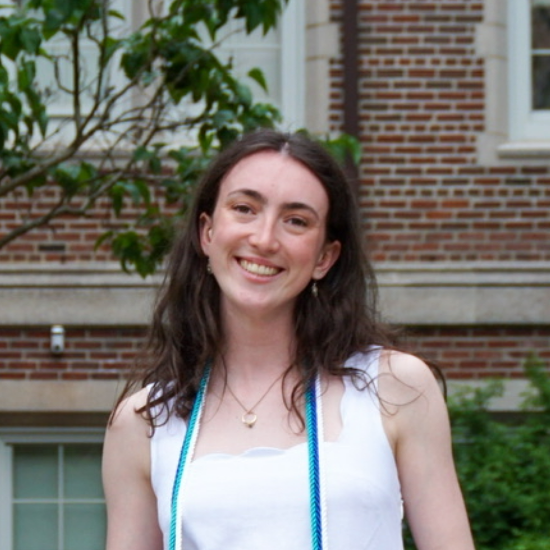 Young woman with long brown hair smiling outdoors in front of a brick building with greenery.