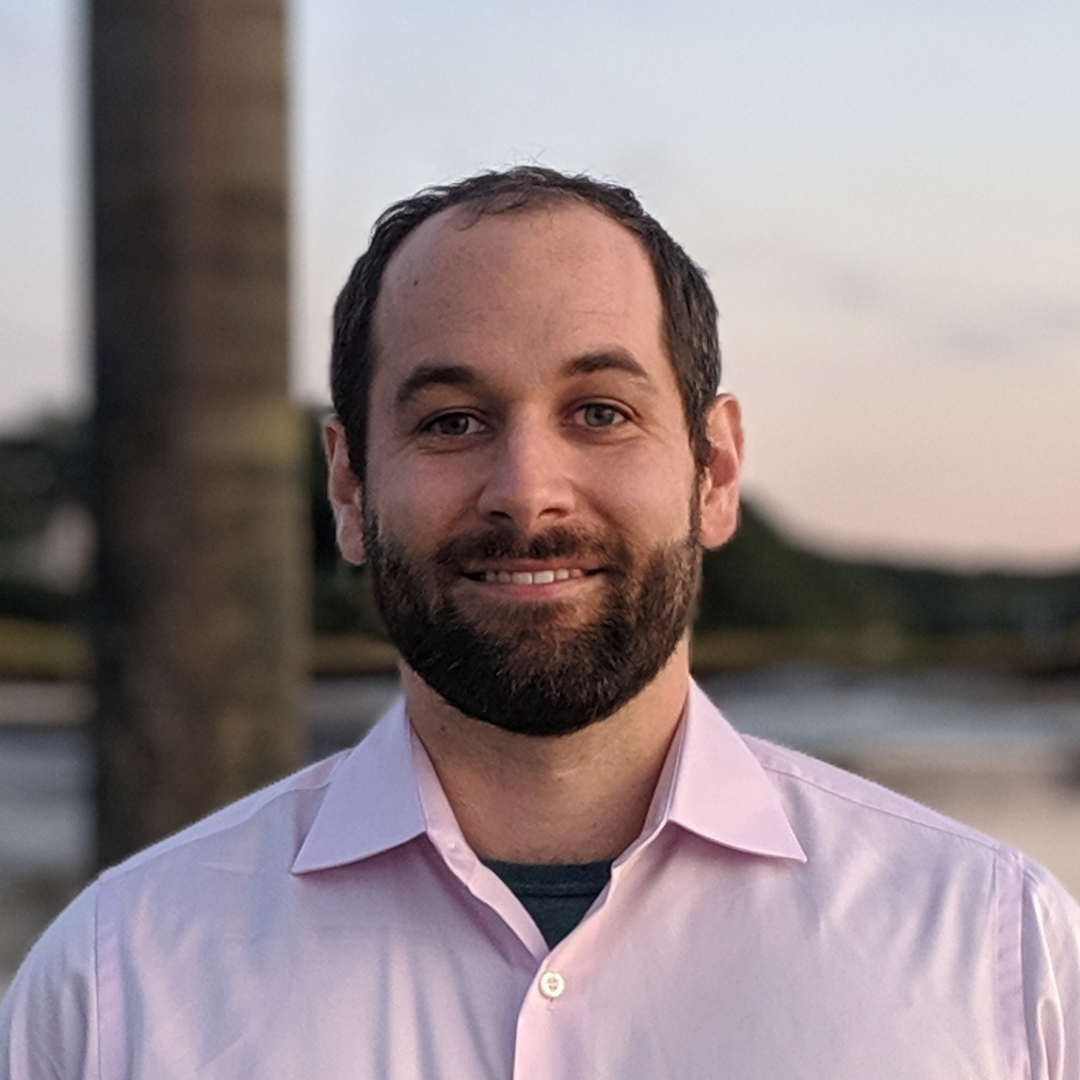 Smiling man with dark hair and beard wearing light pink shirt outdoors near water at dusk.