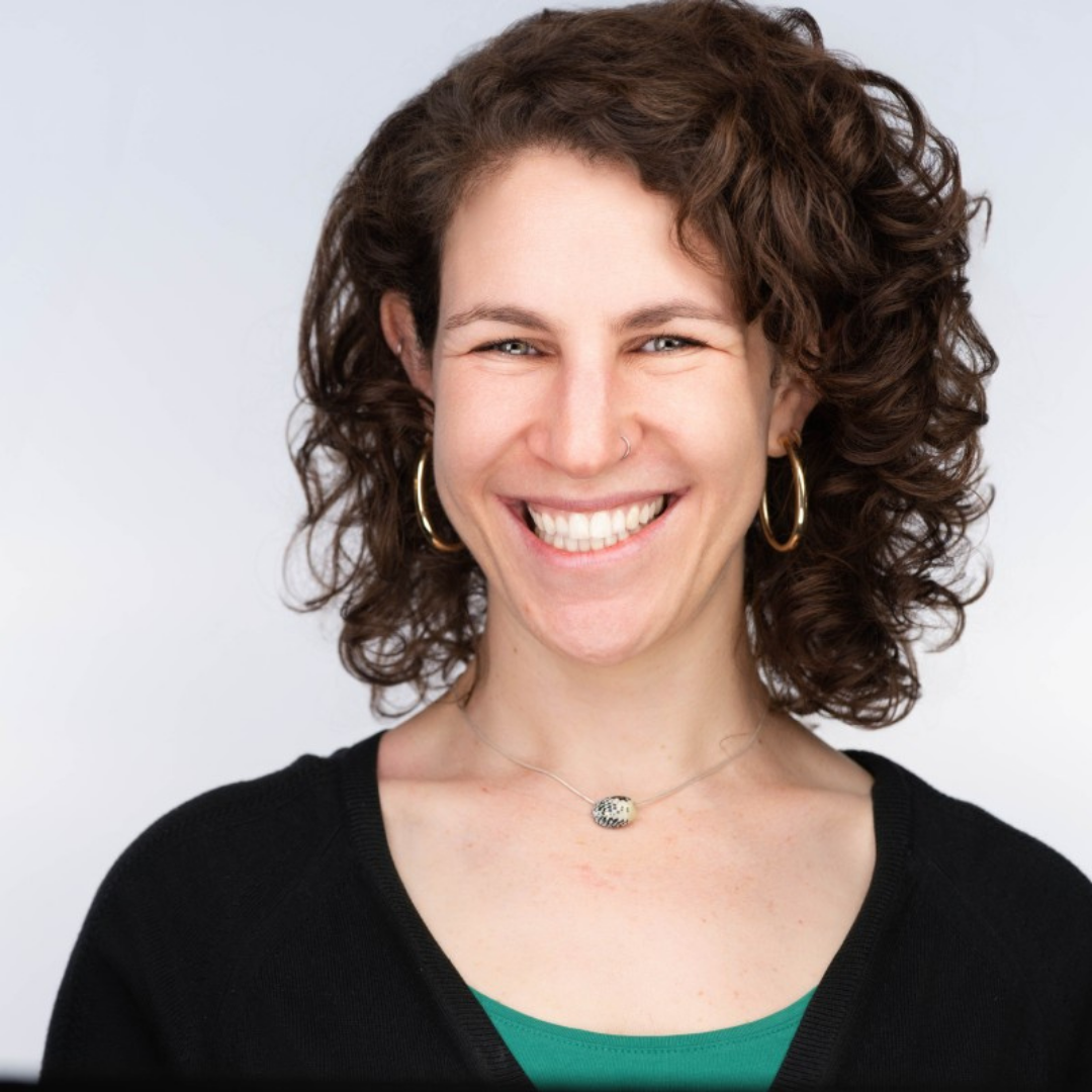 Smiling woman with curly brown hair, wearing gold hoop earrings and a black sweater over a green top.