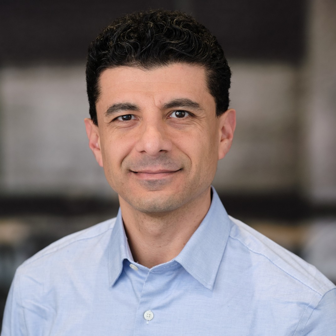 Man with short curly dark hair wearing a light blue button-up shirt against a blurred background.