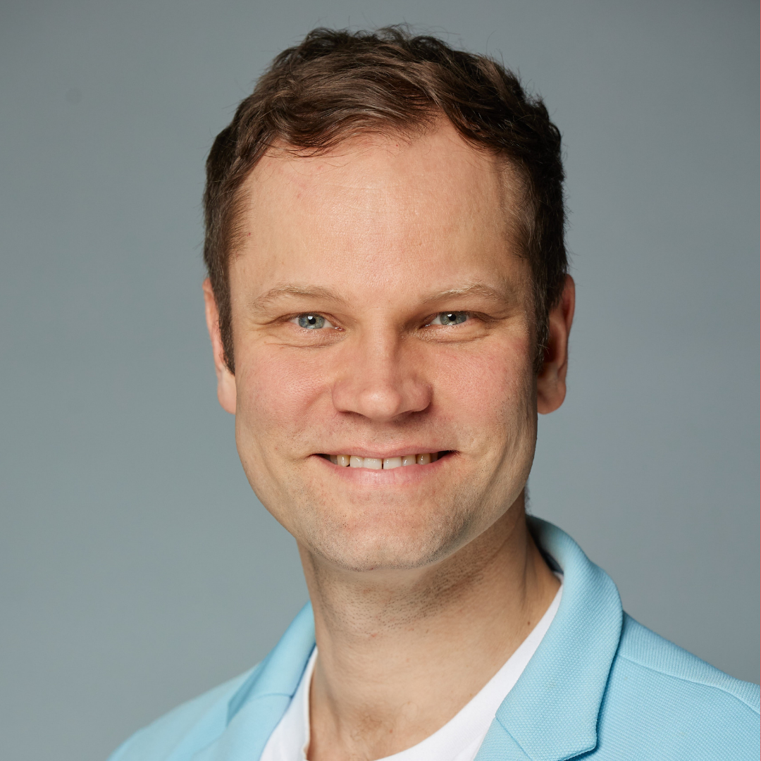 Smiling man with short brown hair wearing a light blue blazer and white shirt against gray background.
