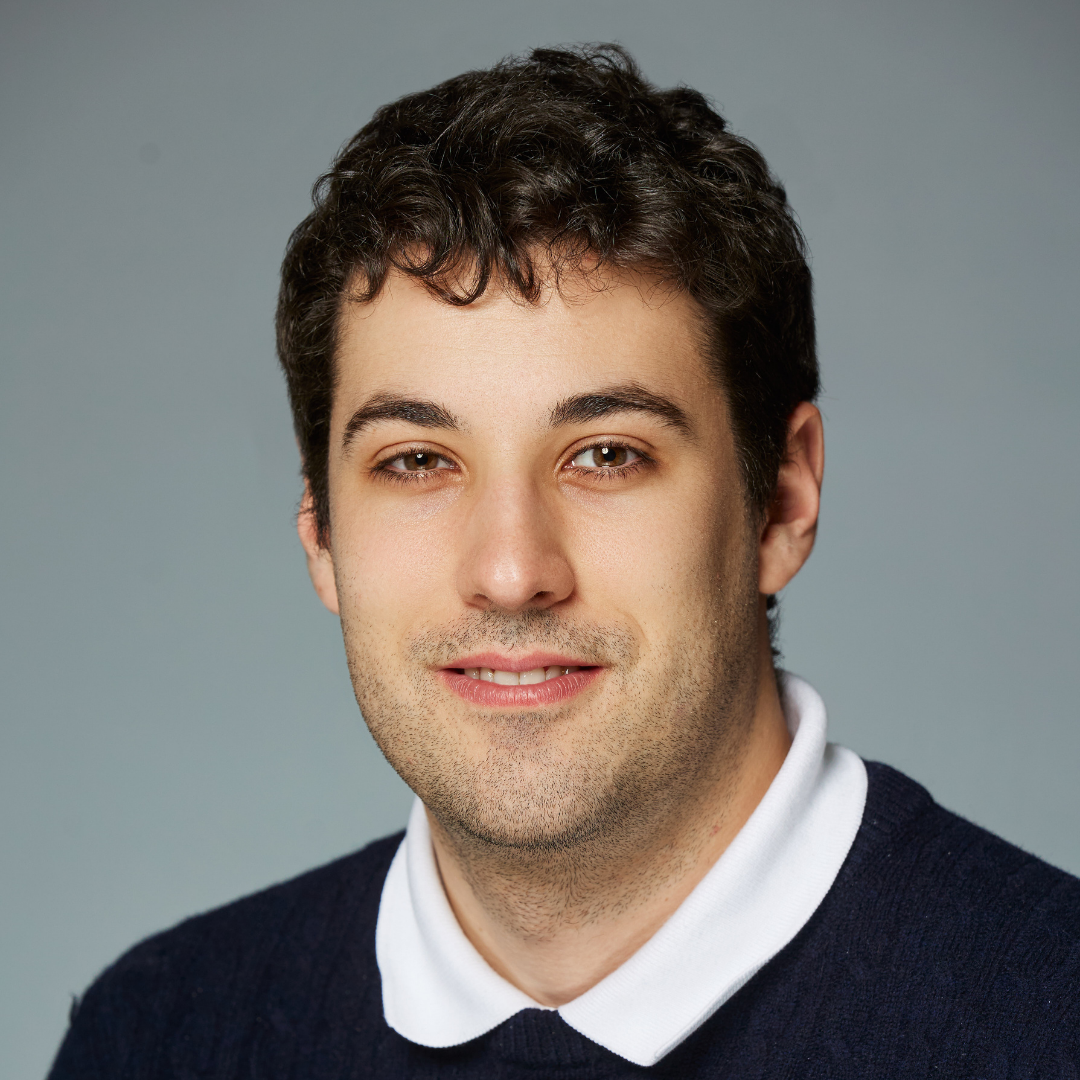 Young man with curly dark hair wearing a navy sweater over a white collared shirt.