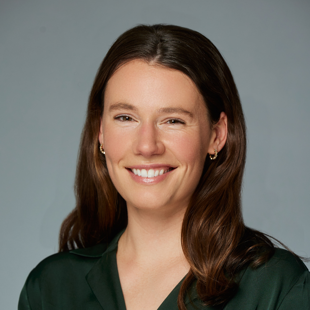 Smiling woman with long brown hair wearing gold earrings and a dark green blouse against a gray background.