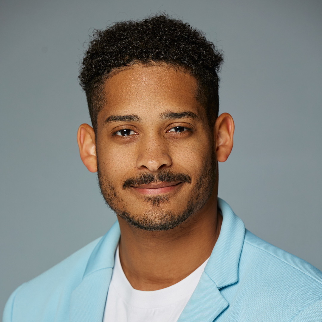 Smiling man with curly hair, light beard, wearing light blue blazer and white shirt on gray background.