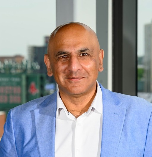 Smiling bald man wearing a light blue blazer and white shirt standing indoors by large windows.
