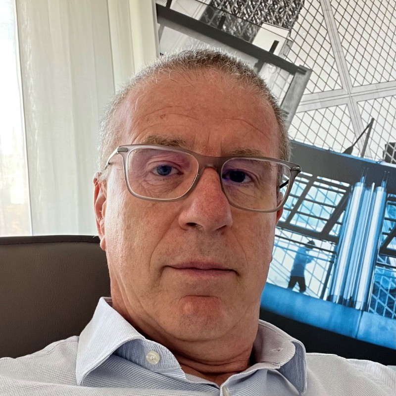 Middle-aged man with glasses wearing a collared shirt, sitting indoors with a geometric patterned background.