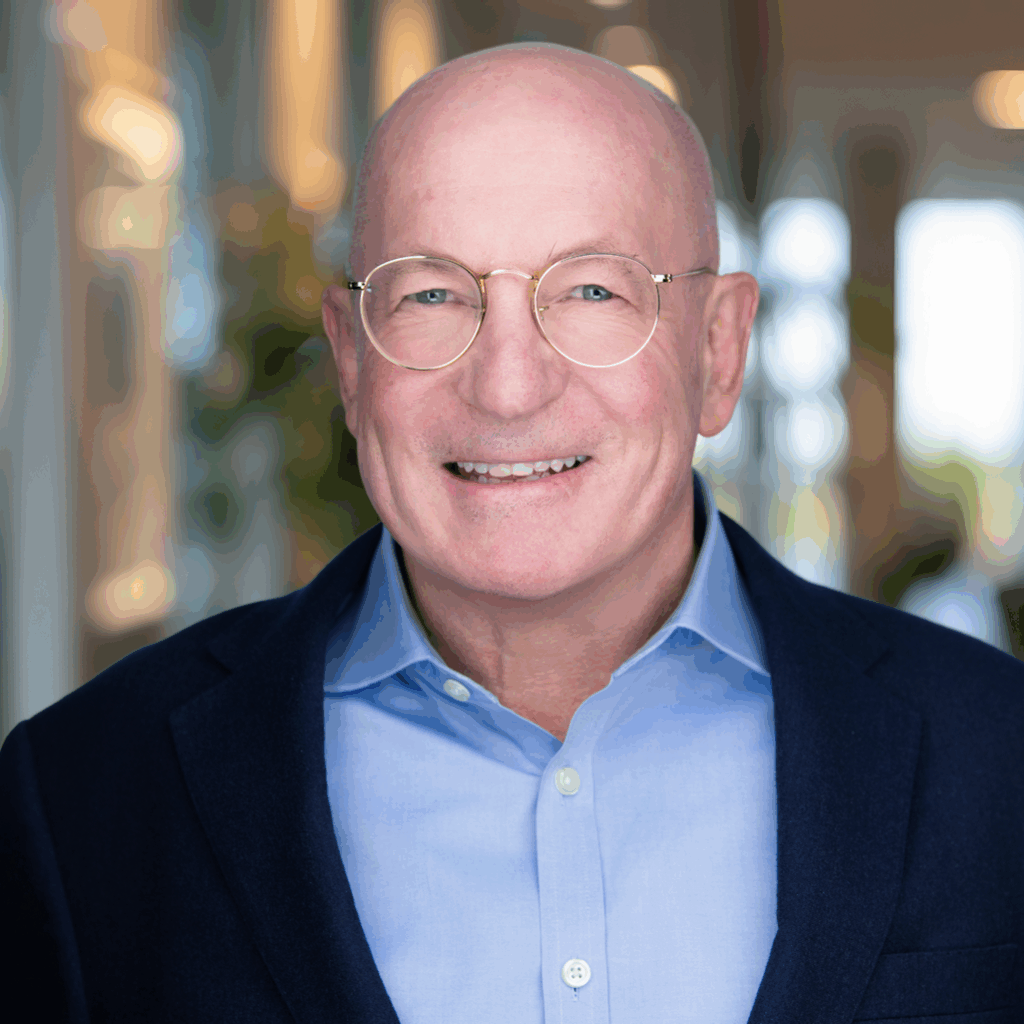 Smiling bald man wearing round glasses, blue shirt, and dark blazer in a warmly lit indoor setting.