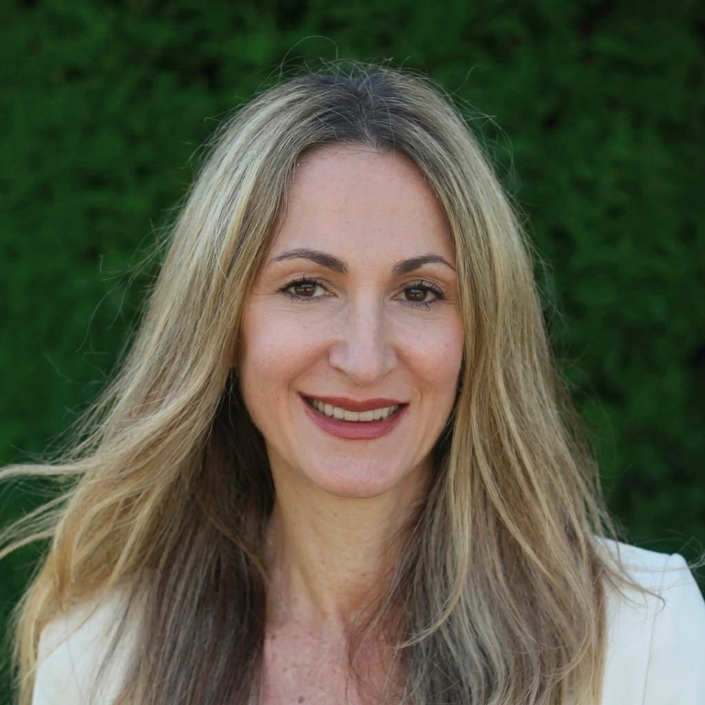 Smiling woman with long blonde hair in a white top against a green leafy background.