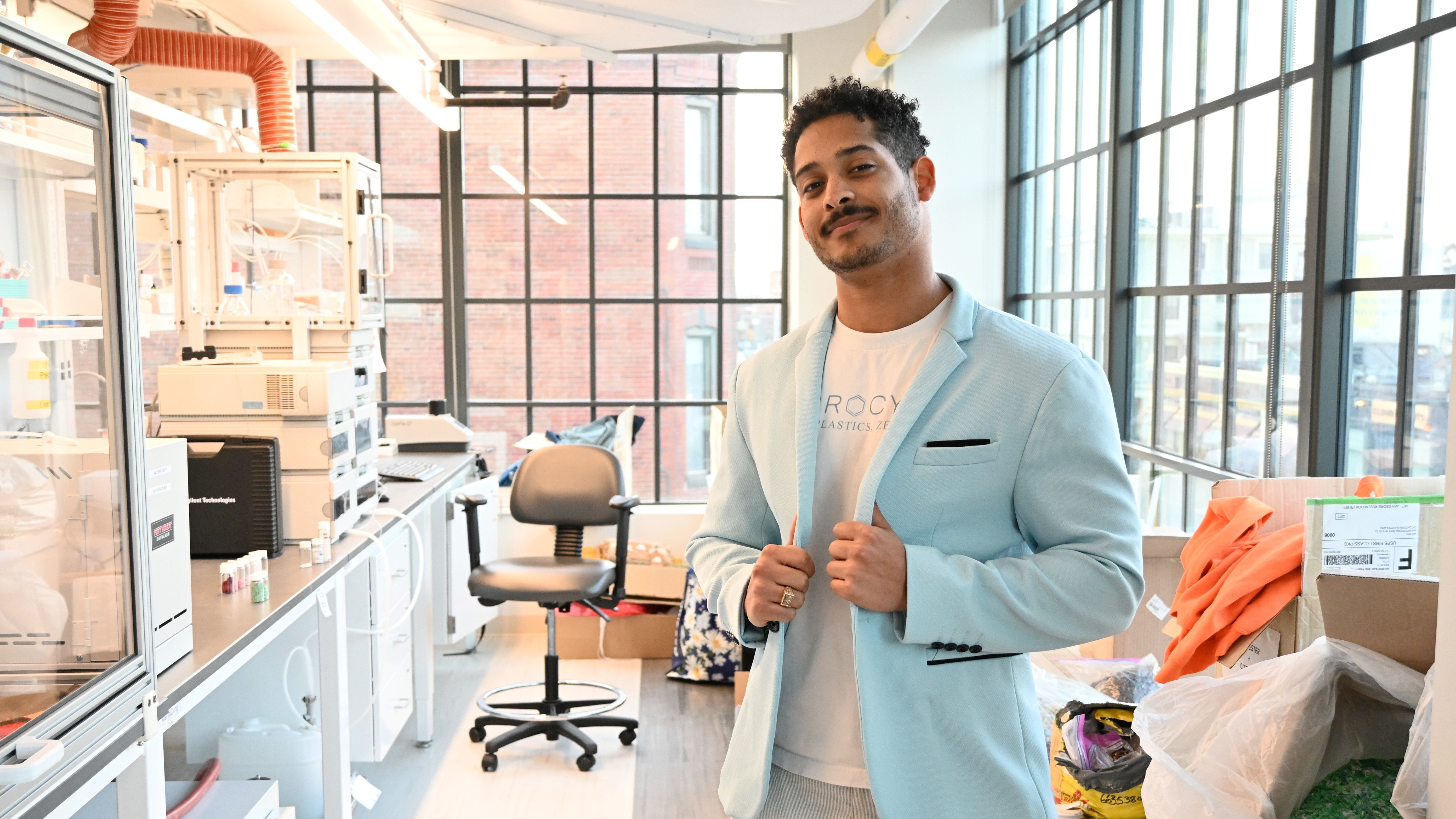 Man in a light blue blazer smiling in a bright lab with large windows and scattered supplies.