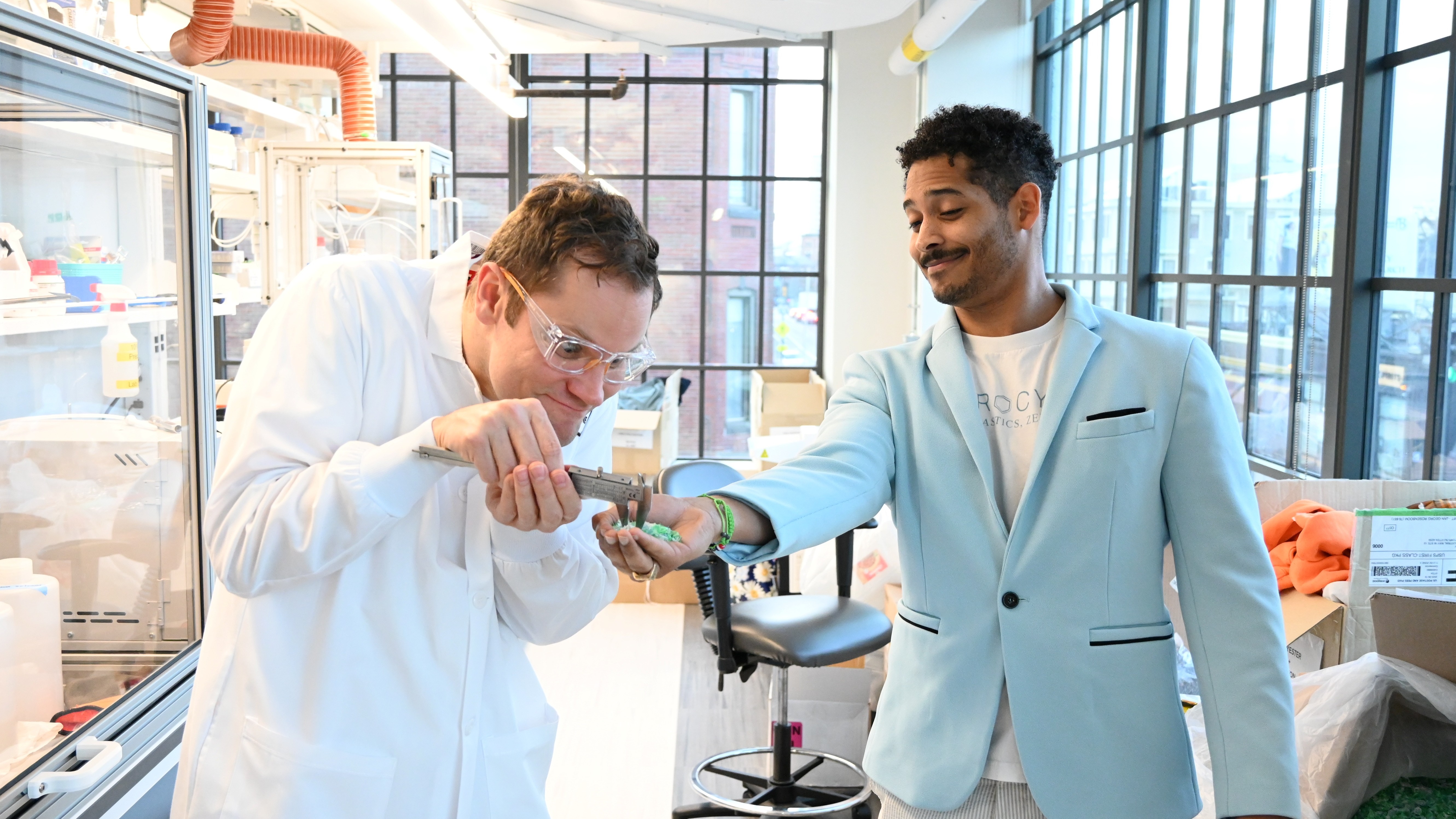 Scientist in lab coat measures green material in another man's hand in bright lab with large windows.