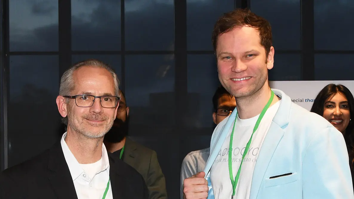 Two men smiling, one in glasses and a black jacket, the other in a light blue blazer and white shirt.