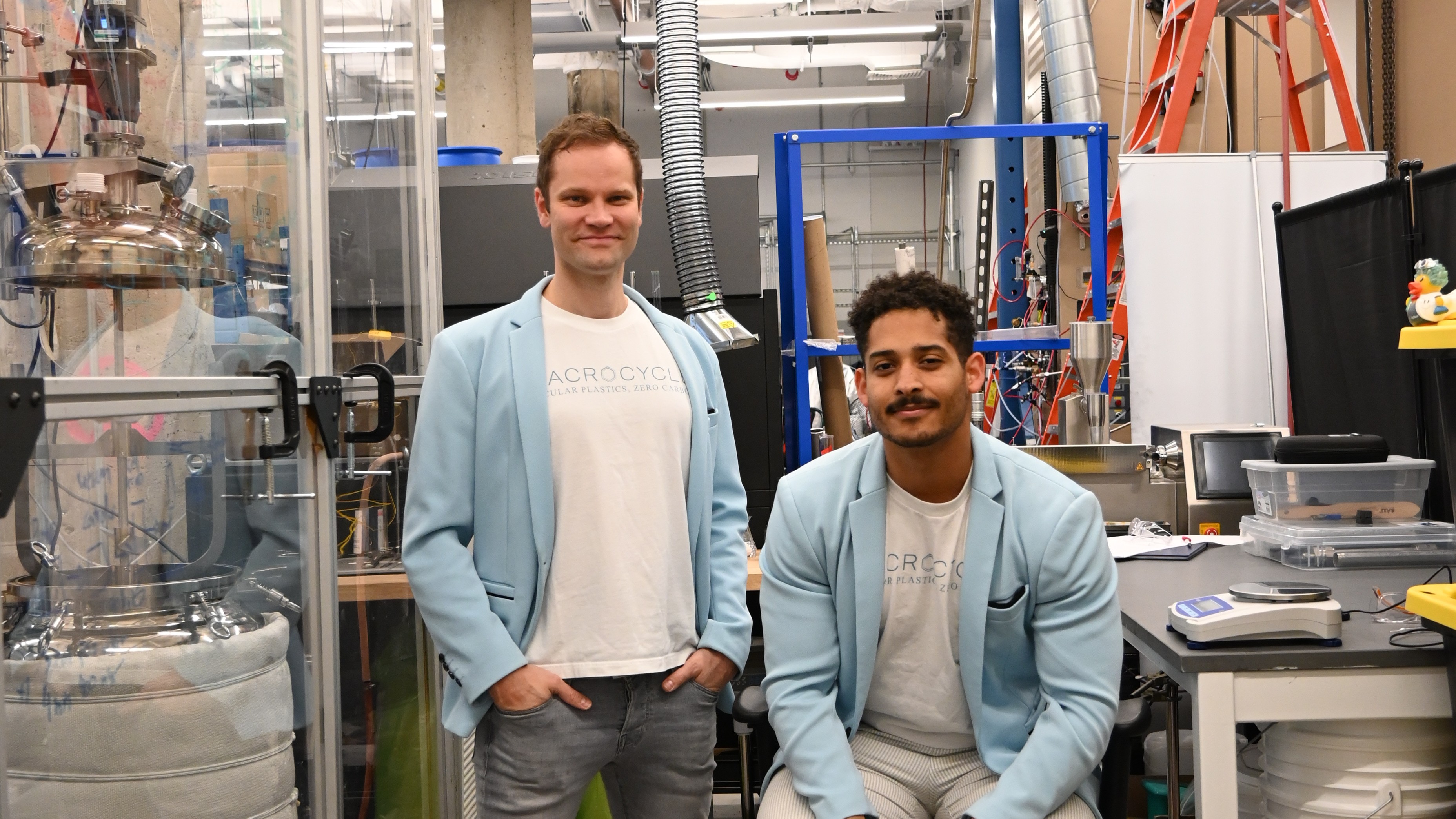 Two men in light blue blazers and white shirts pose in a lab with scientific equipment around them.