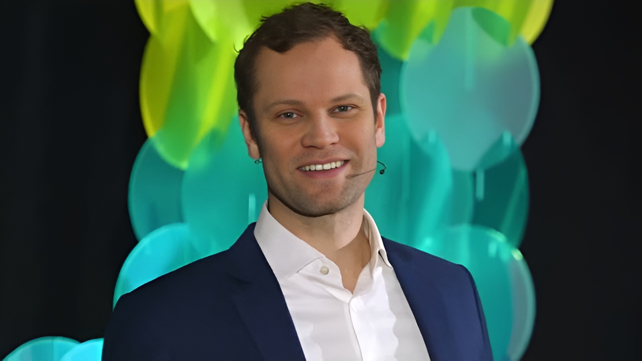 Smiling man in a dark blue jacket and white shirt speaking with a headset microphone and colorful round background.