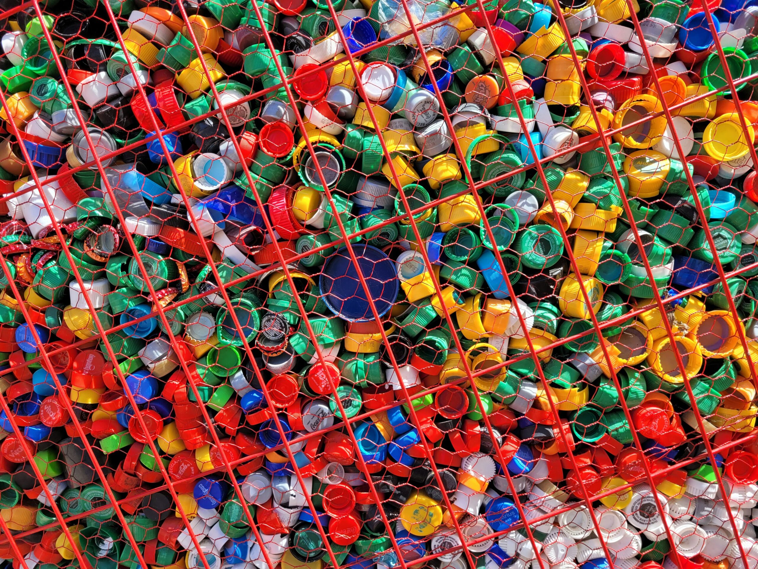 Pile of assorted colorful plastic bottle caps contained by red wire mesh.