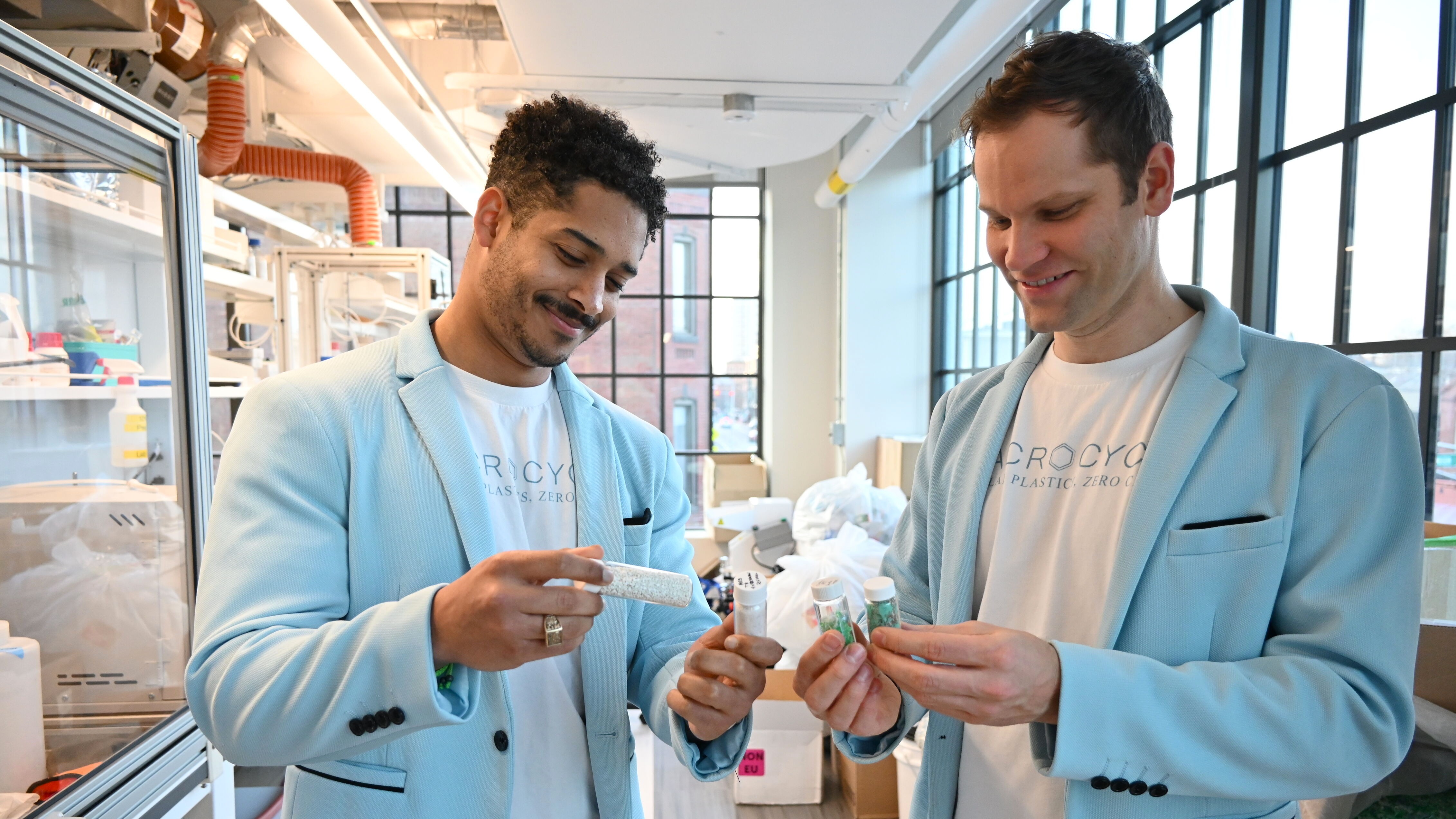 Two men in light blue blazers examining clear vials with materials inside in a bright workspace.