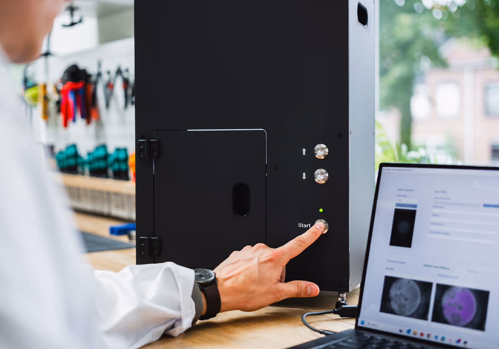 Person pressing start button on black scientific device connected to laptop with data on screen.