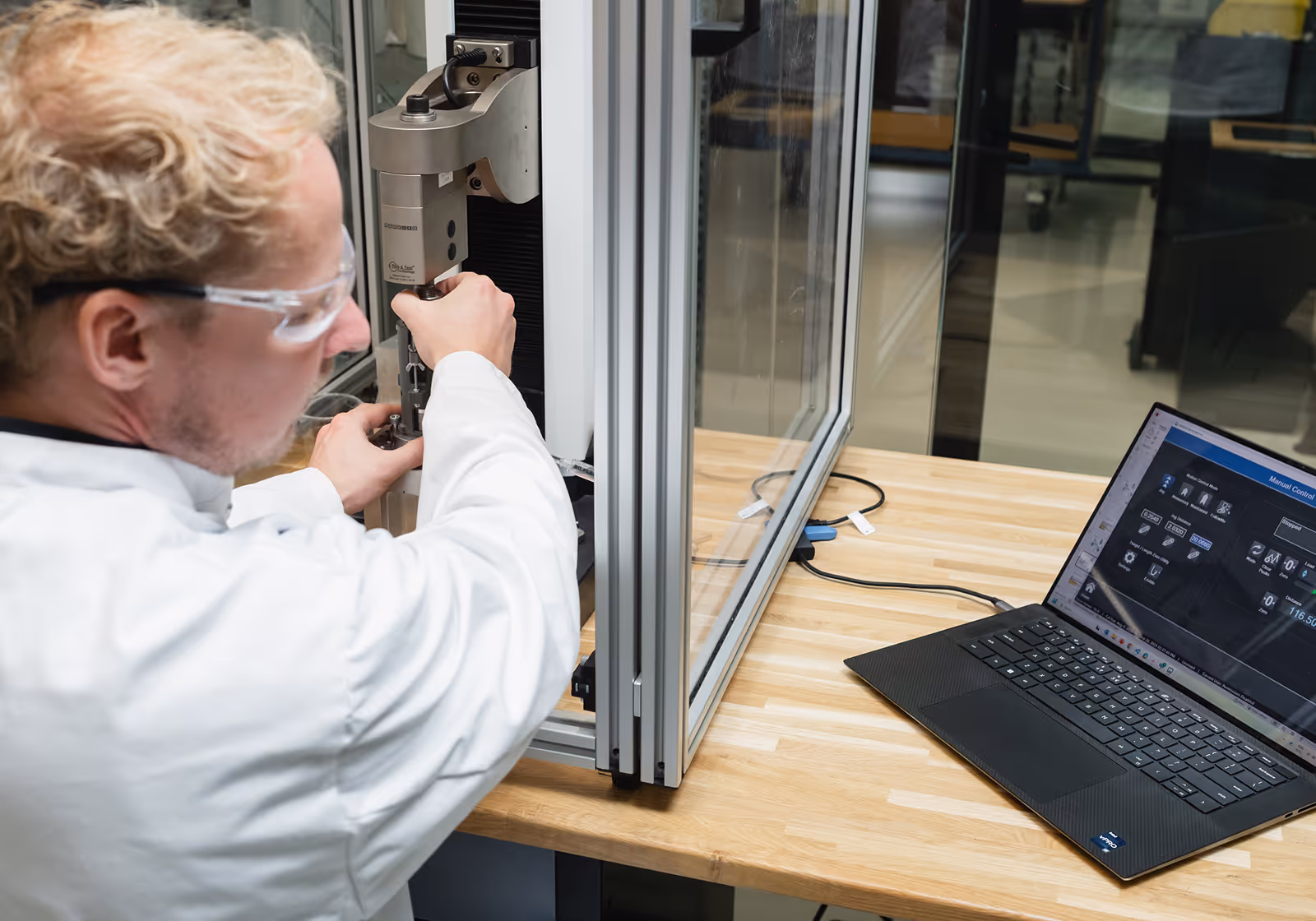 Scientist in lab coat adjusting a machine next to a laptop with control software on screen.