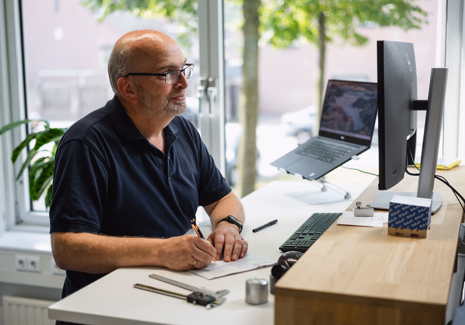Older man with glasses writing on paper at desk with computer monitor and laptop near window.