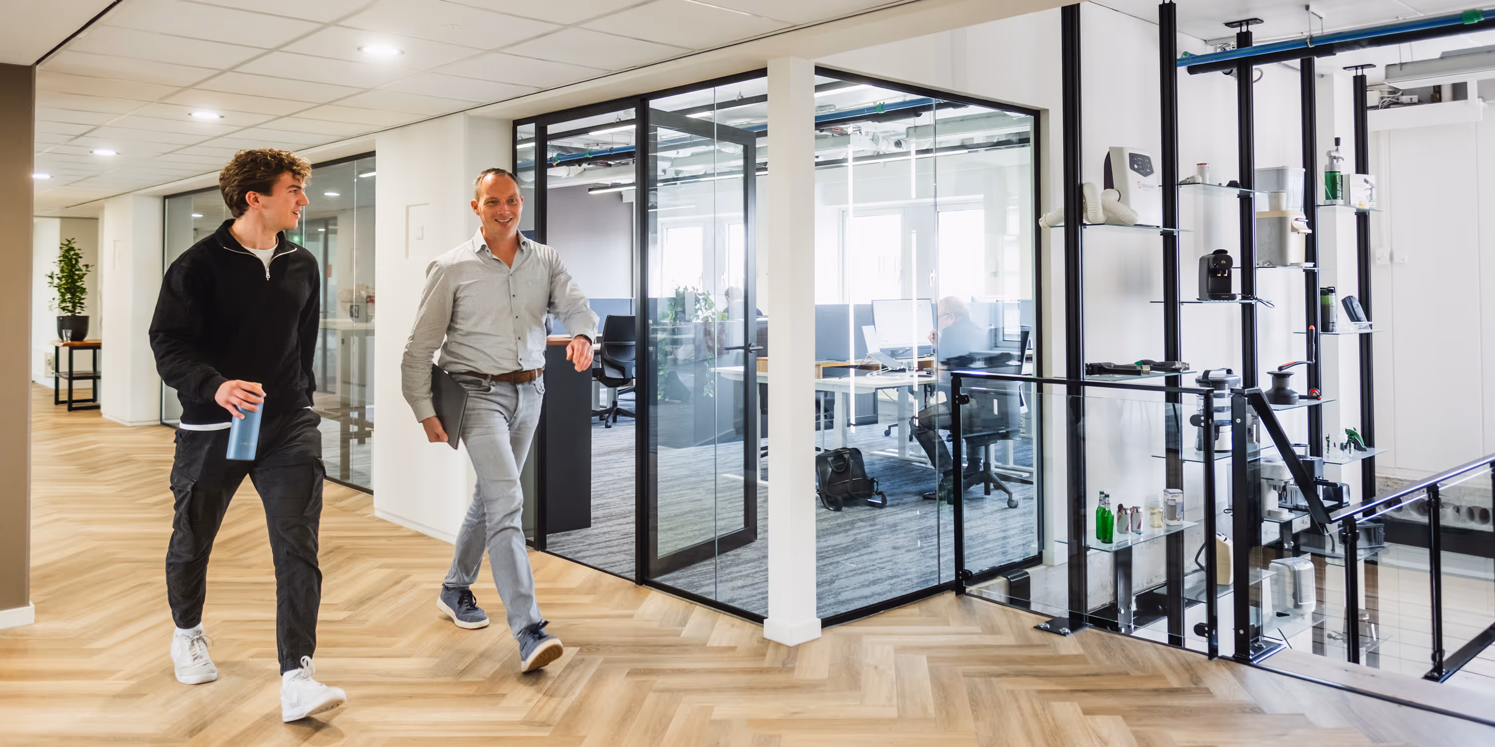 Two men walking and talking in a modern office with glass-walled rooms and wooden floors.