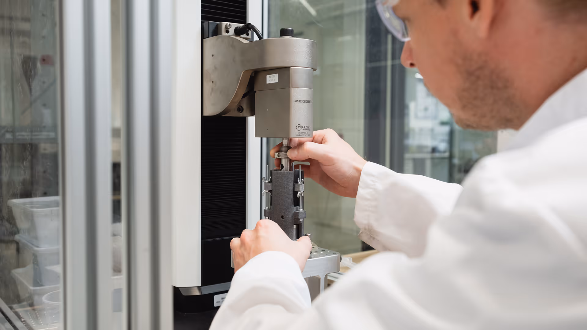 Scientist in lab coat and safety glasses adjusting a machine for material testing in a lab.