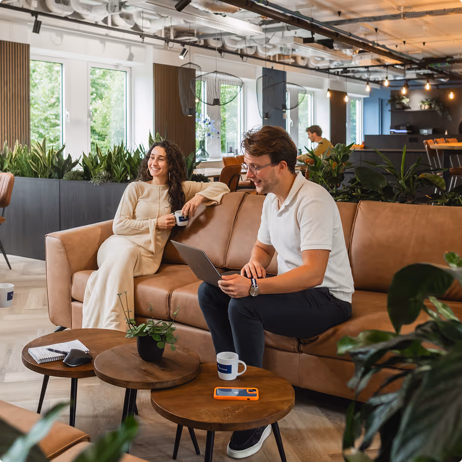 Two people smiling and talking on a brown couch in a modern, plant-filled office lounge.