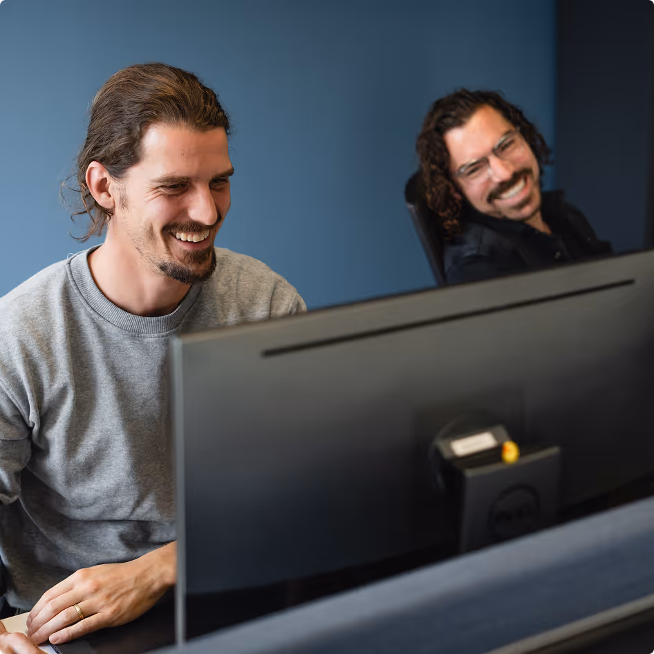 Two men smiling and looking at a computer screen together in an office setting.