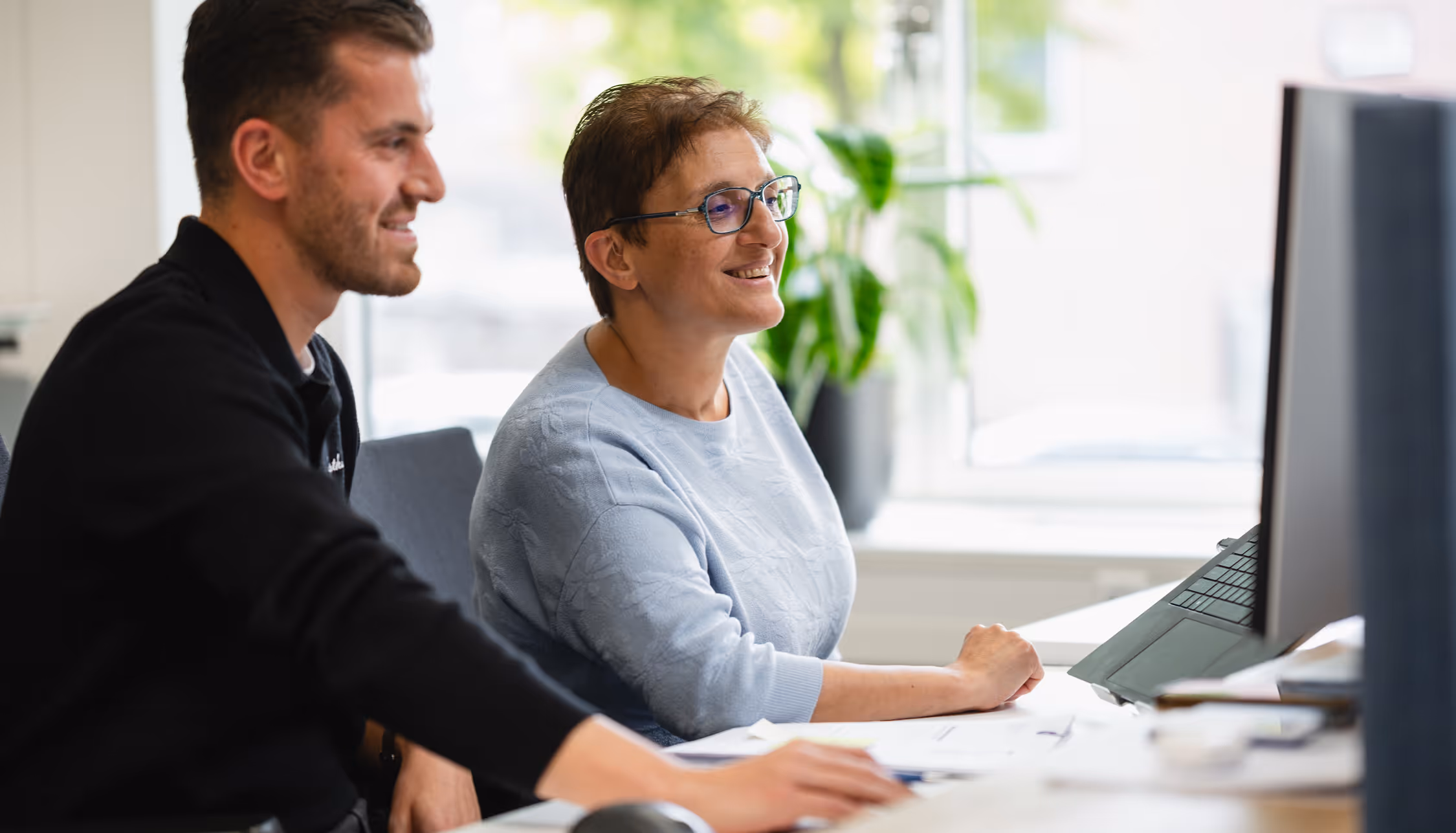 Two colleagues smiling and working together at a computer in a bright office with a green plant by the window.