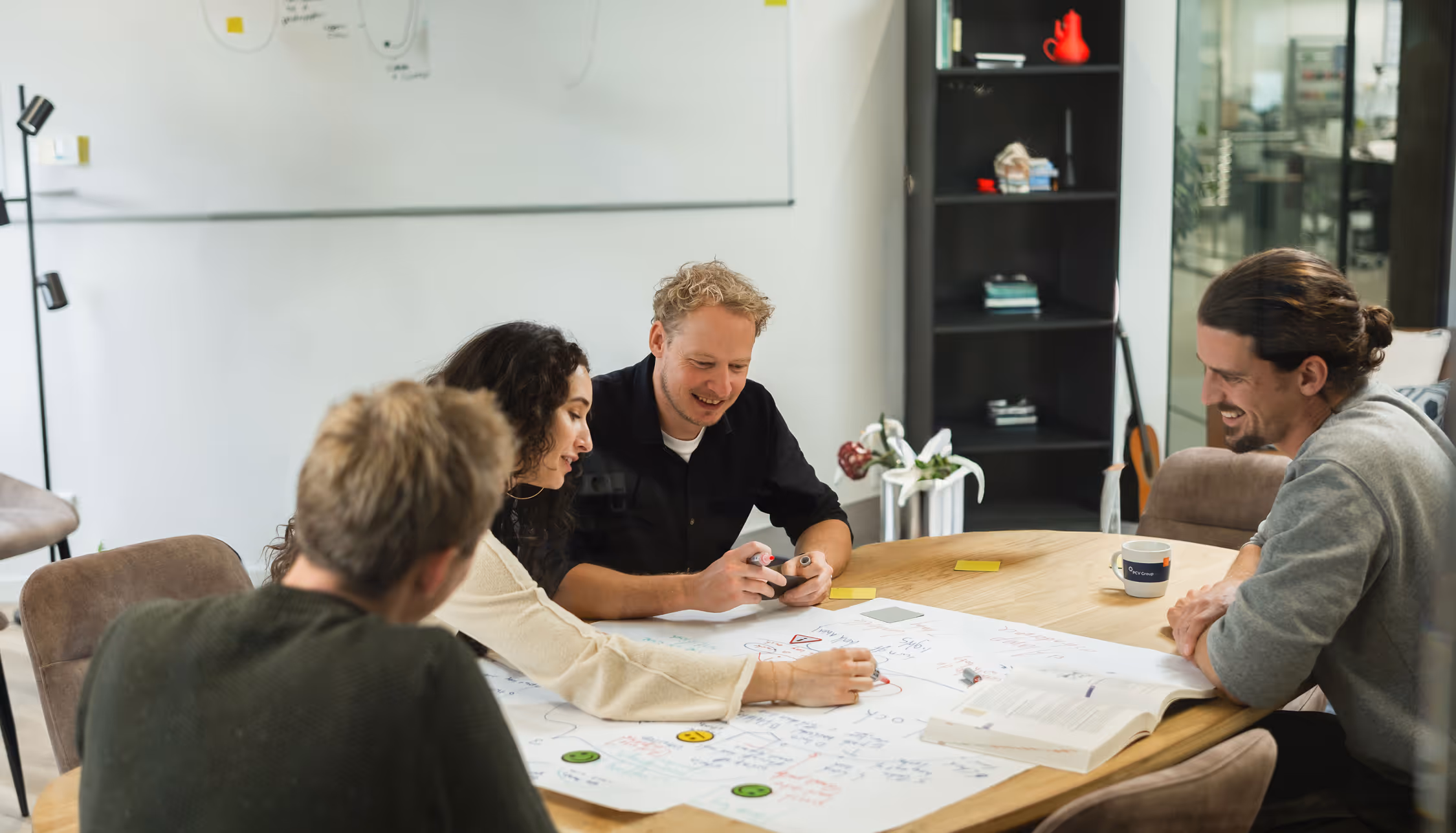 Four people sitting around table brainstorming on large paper with notes and markers in office.