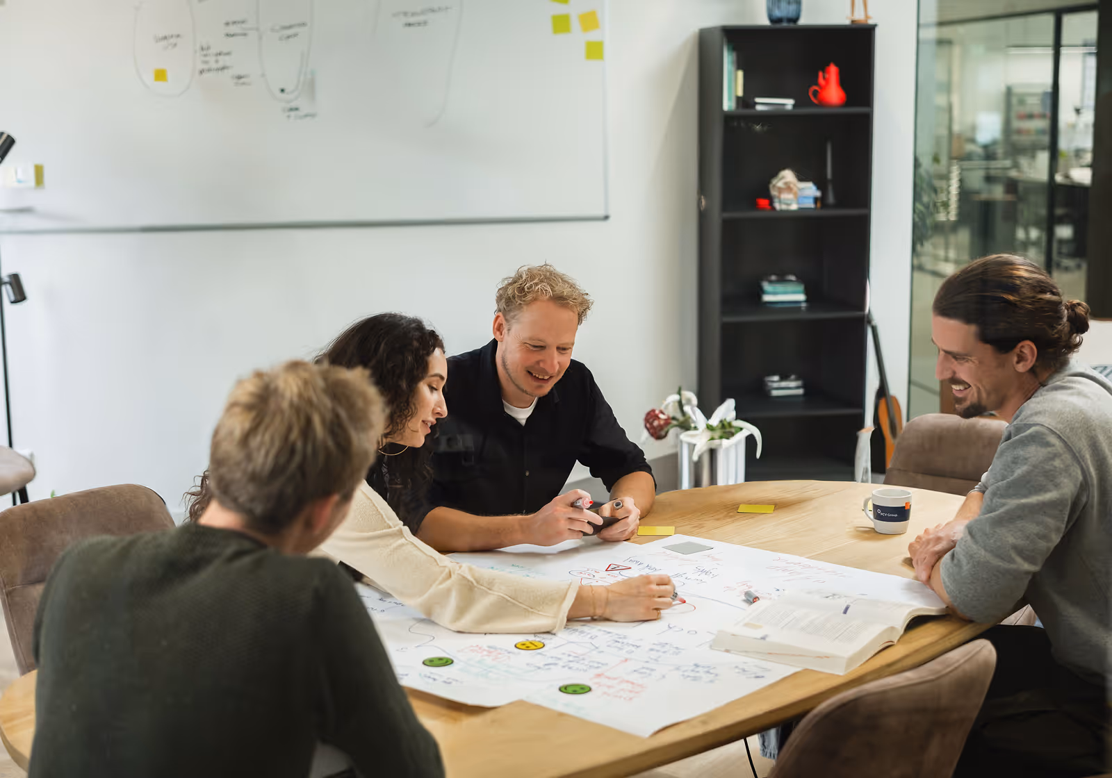 Four people collaborate around a table with charts, notes, and an open book in a modern office.