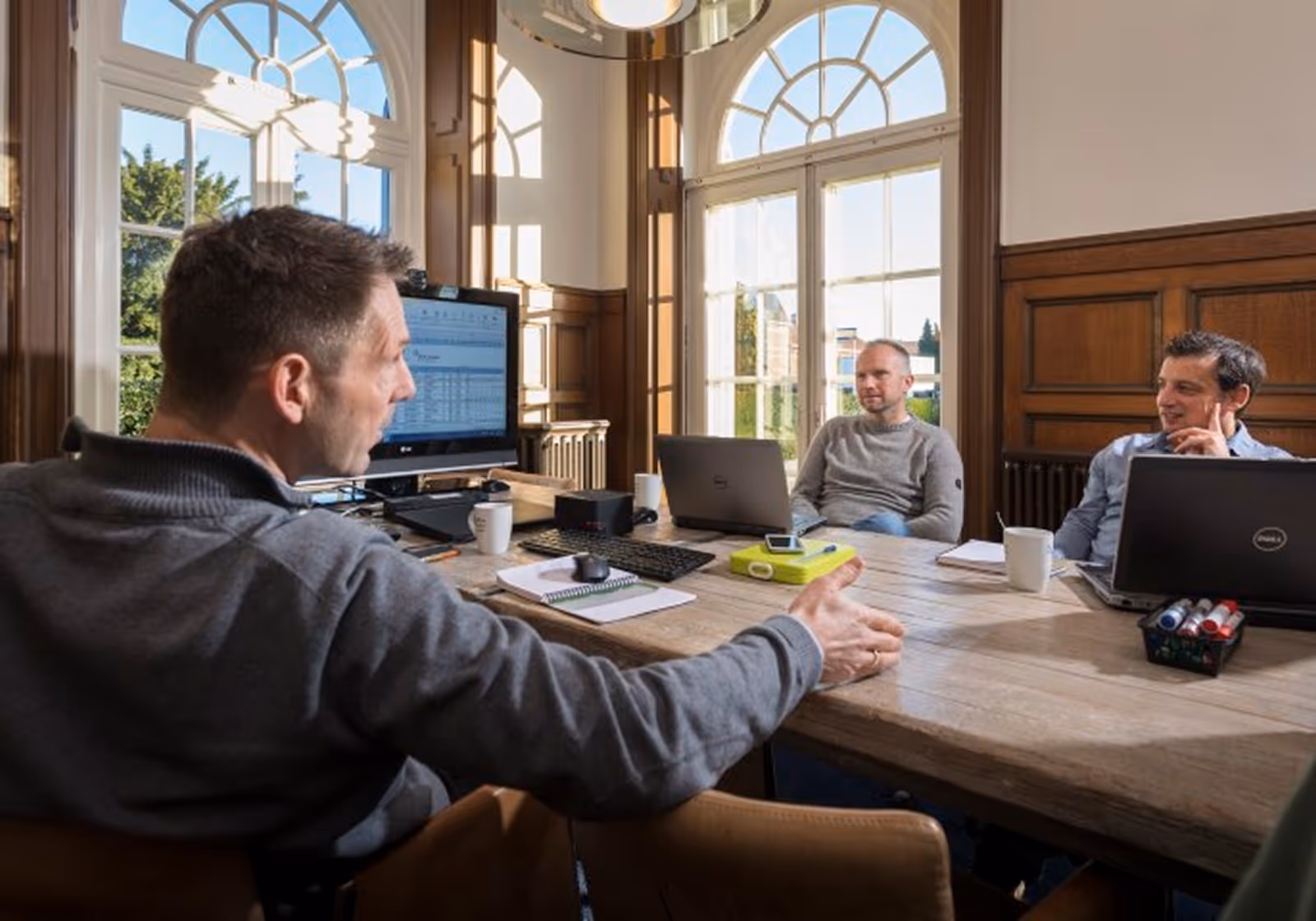 Three men having a discussion at a wooden table in a bright office with laptops and papers.