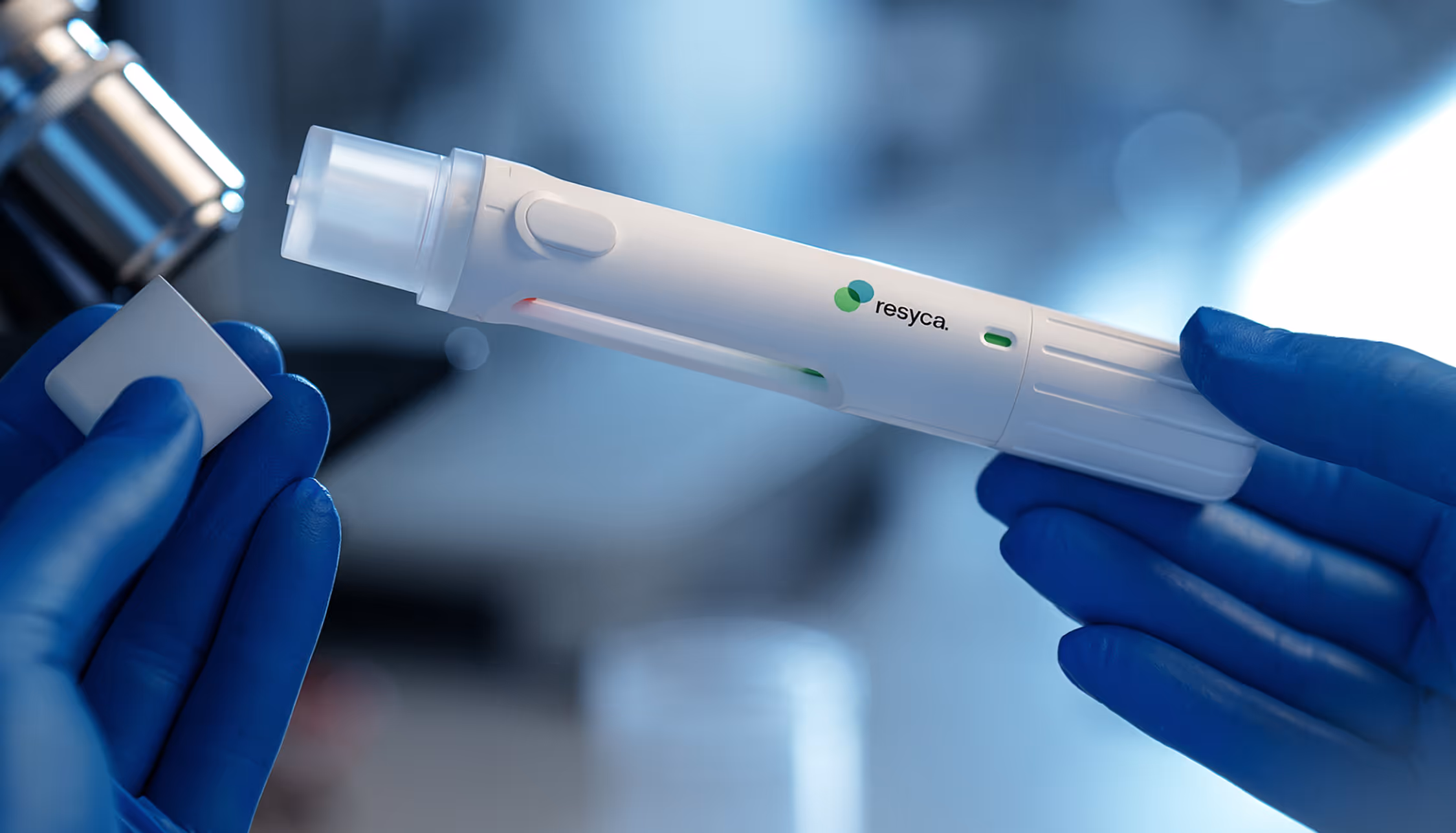 Gloved hands holding a Resyca-branded medical device near a microscope in a lab setting.