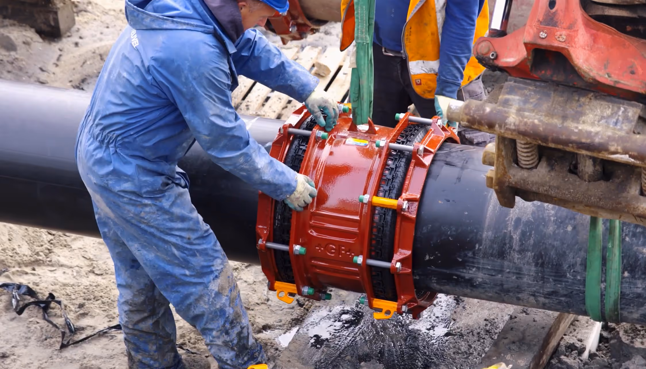 Workers installing a large red mechanical coupling on a black pipeline at a construction site.