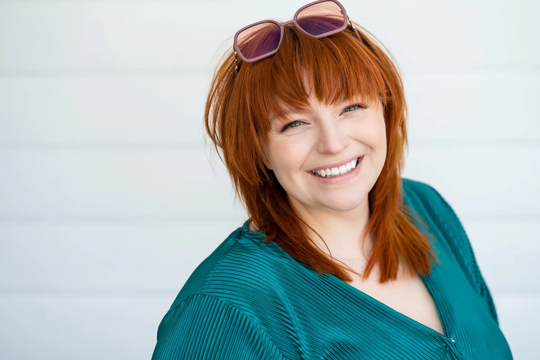 A woman laughing against a white wall, sunglasses pushed up in her hair