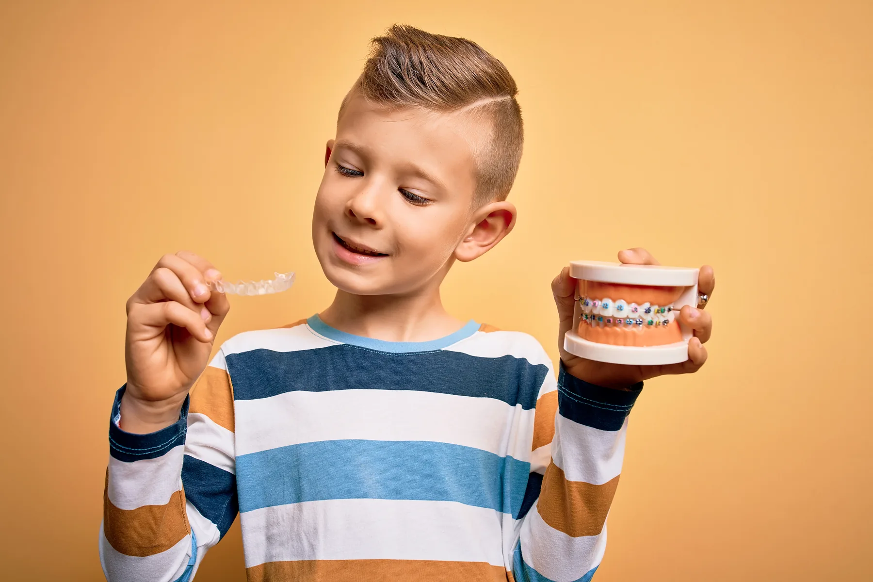 A boy holding a clear aligner in one hand and a model of braces in the other