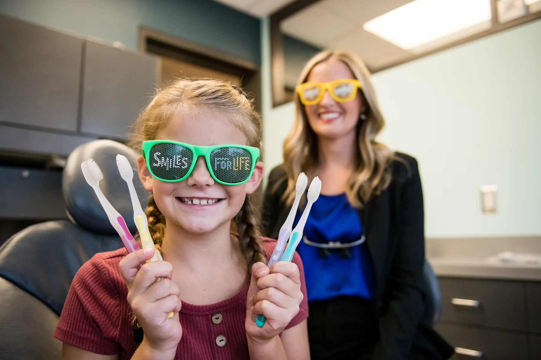 A girl in novelty sunglasses holding up new toothbrushes, a team member laughing behind her