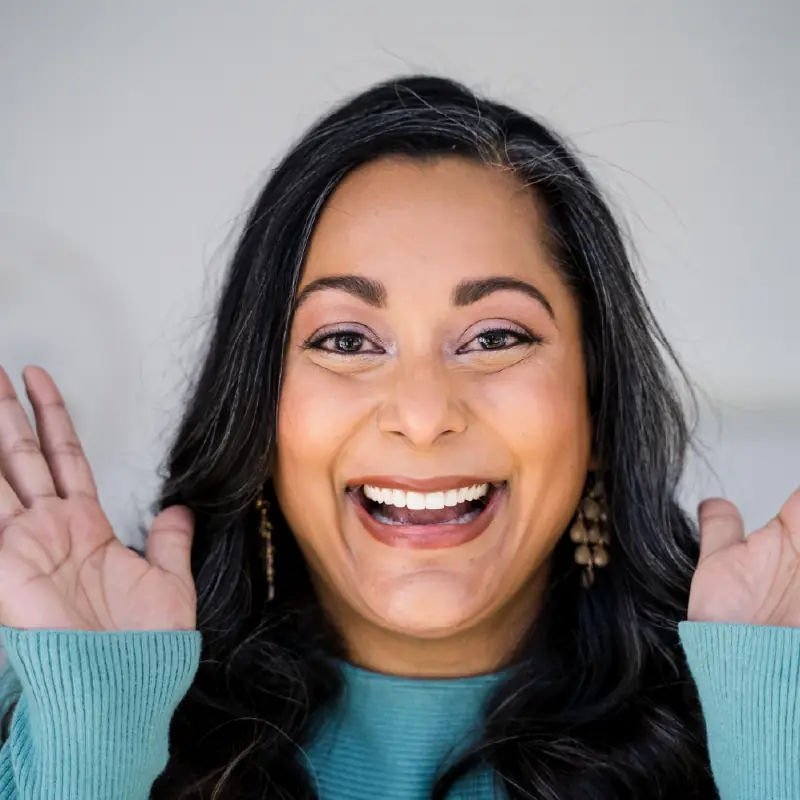 Smiling Redmond Way Dentistry patient with long dark hair wearing teal sweater, holding hands up by face against gray background.