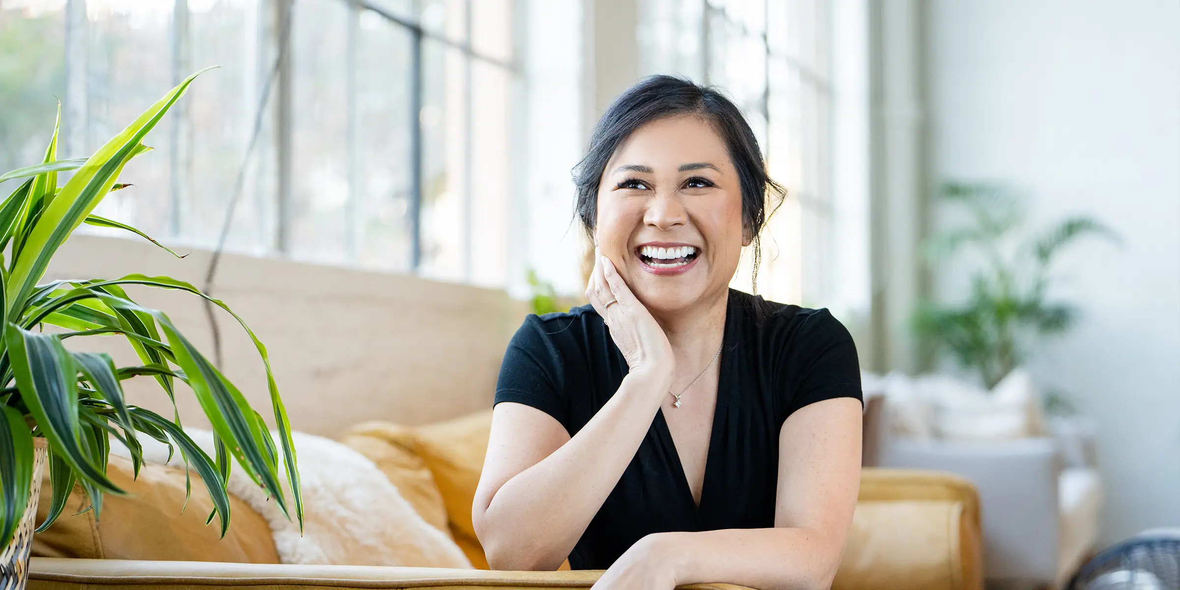 Smiling woman with black hair sitting on a couch in a bright room with plants around her.