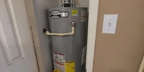 Gray water heater tank installed in a small corner space near a door and a light switch on a beige wall.