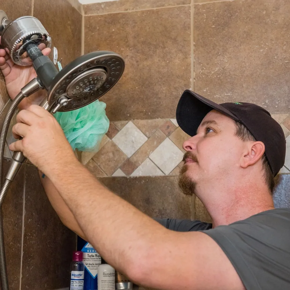 Man in a black cap installing or repairing a showerhead in a tiled bathroom.