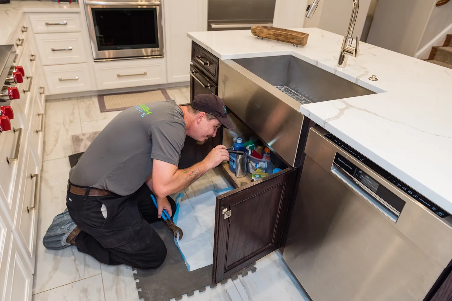 Plumber kneeling in kitchen inspecting under sink cabinet with a flashlight and wrench.