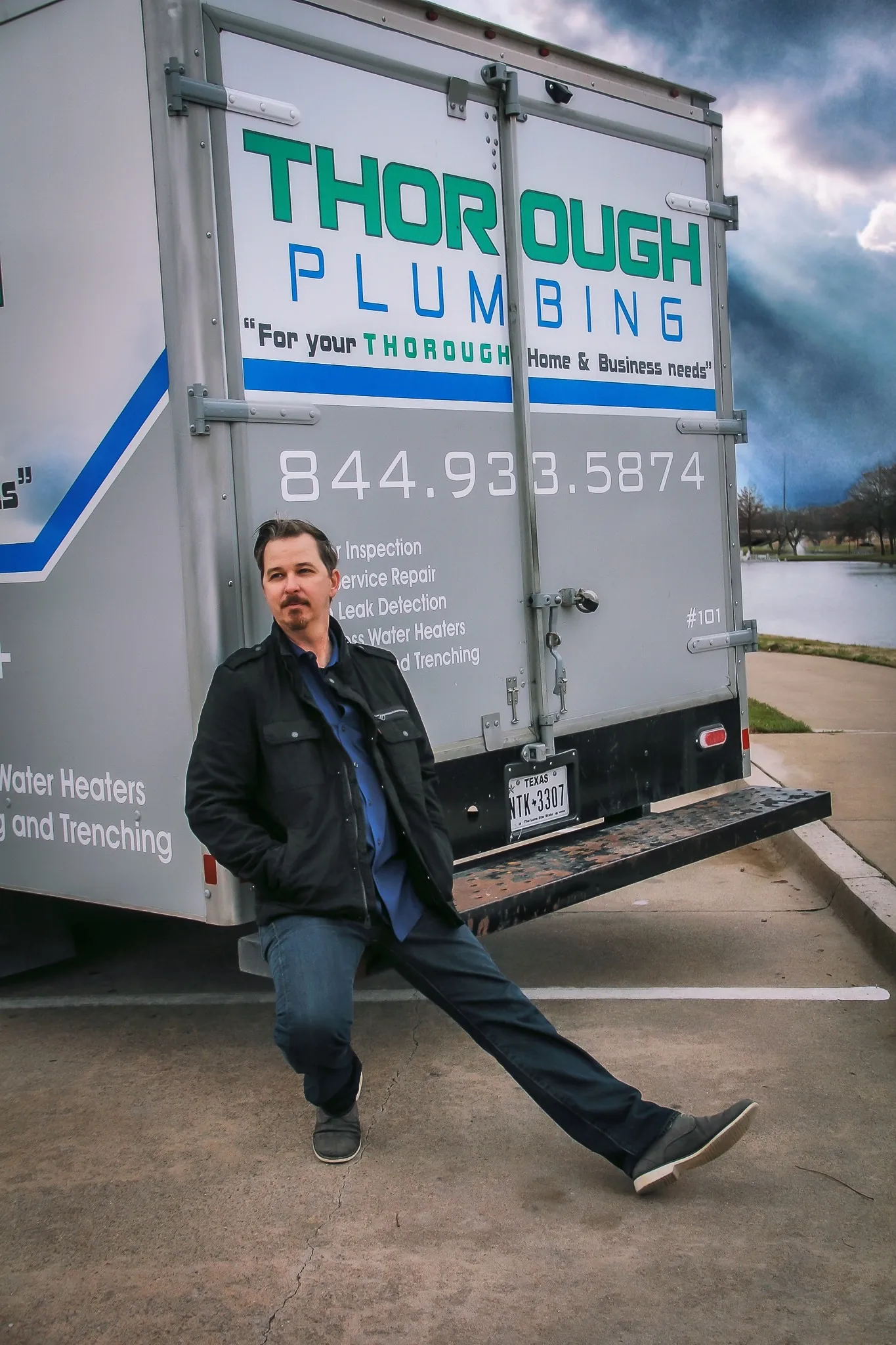 Man in black jacket and blue shirt sitting with legs extended beside a plumbing service truck.