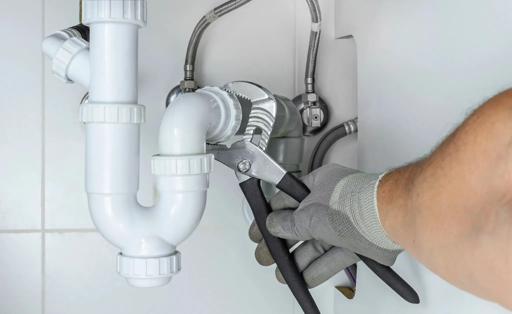 Gloved hand using pliers to tighten a white plastic pipe joint under a sink.