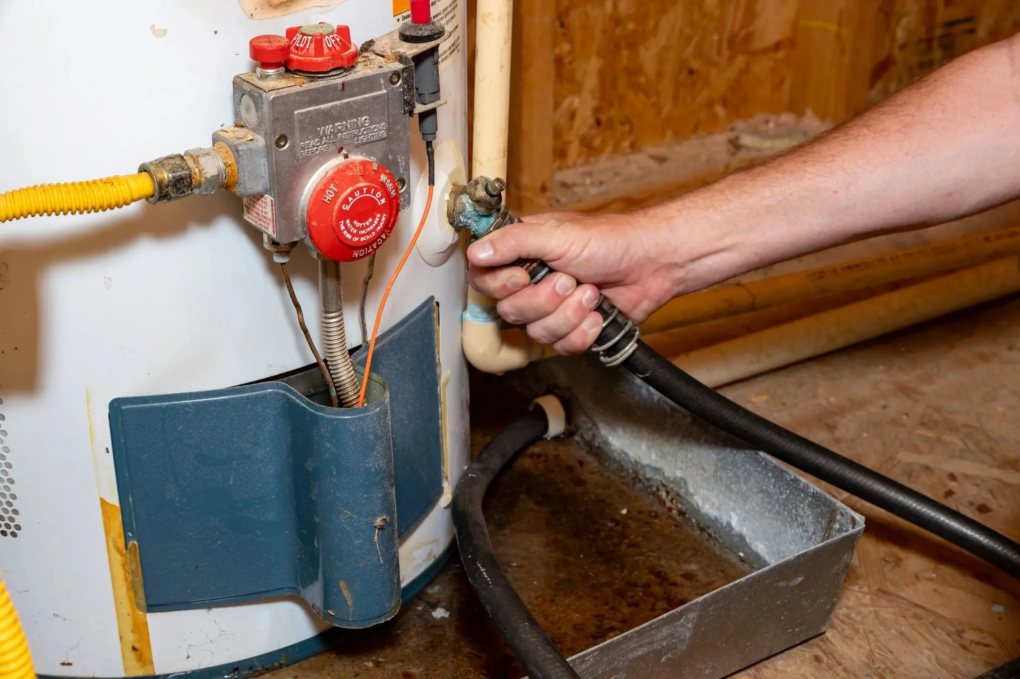 Hand holding hose connected to valve on a water heater with a drip pan underneath.