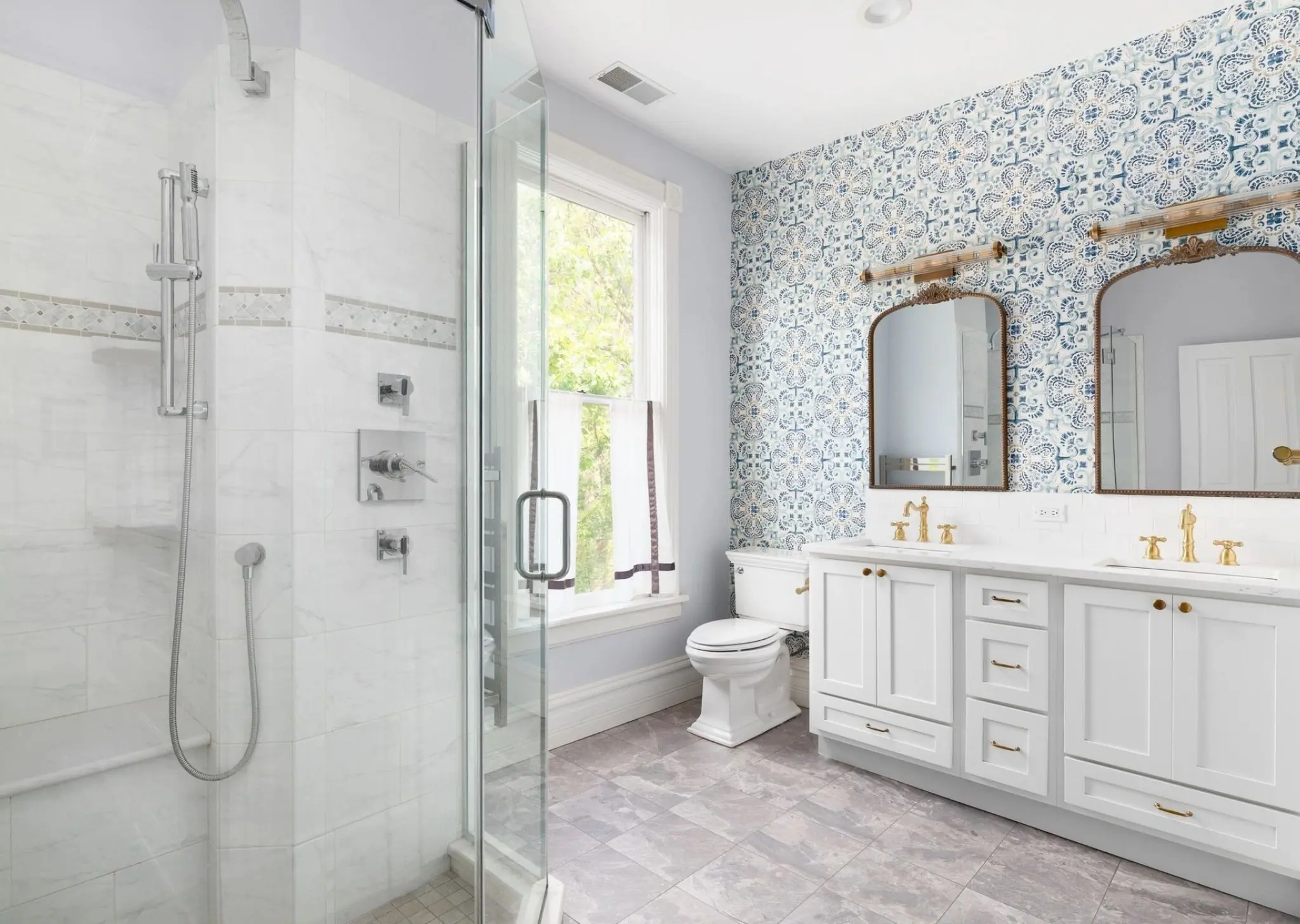 Bright bathroom with glass shower, floral tiled wall, double sinks with gold fixtures, and a white toilet.