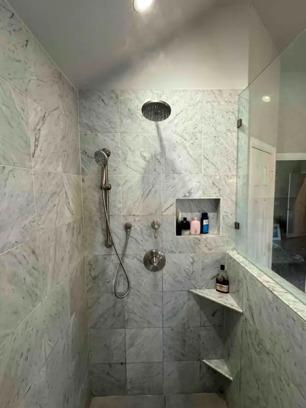 Marble-tiled shower with rainfall and handheld showerheads, built-in shelf with toiletries, and corner shelves.